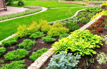 A landscaped garden with neatly arranged shrubs and greenery, bordered by stone edging, and a well-manicured lawn in the background.